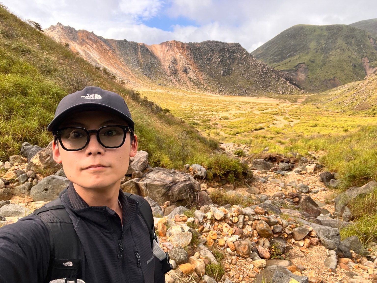 午後の晴れ間に広い草地と山の斜面を背景に撮った筆者の写真